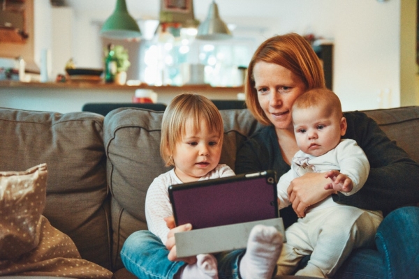 Red-haired mother holds a baby on her lap while a young child next to her touches a tablet computer